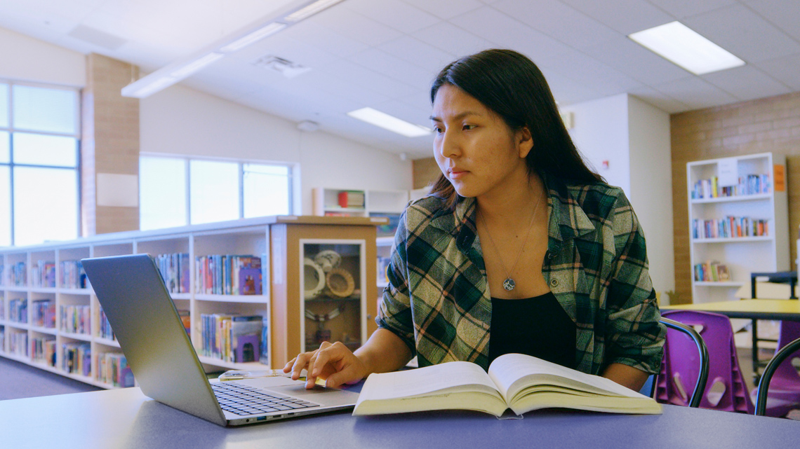 Student with an open book at a desk and in front of a portable computer.