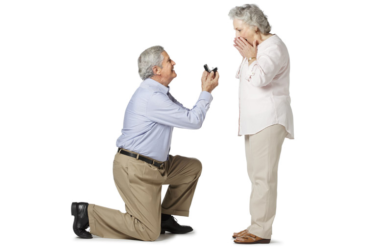 Elderly man down on one knee with a jeweler’s box open facing a surprised elderly woman