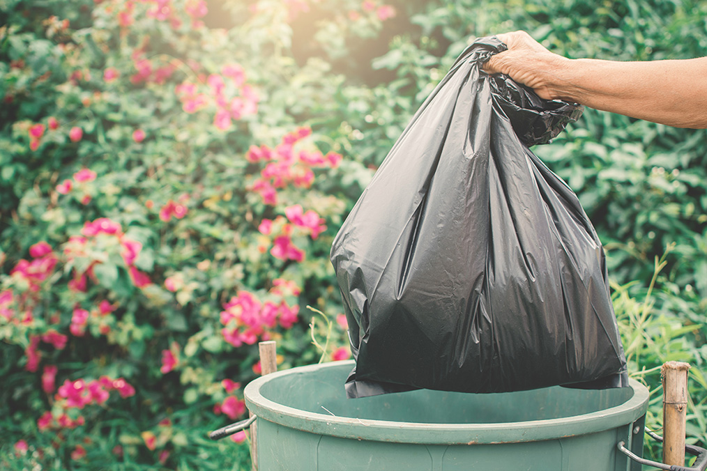 Main d’une personne âgée jetant un sac plastique plein dans une poubelle en plastique.
