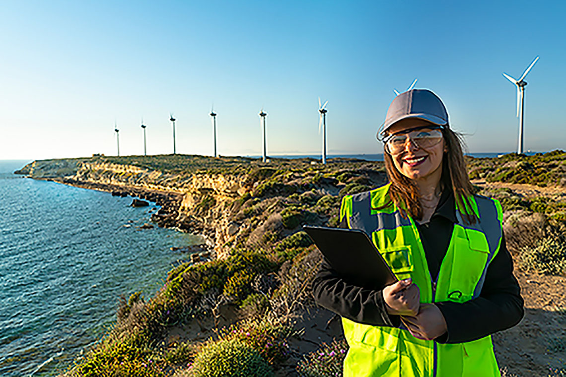 Person with reflective vest & safety glasses, holding a tablet. Background of windmills on a shore.