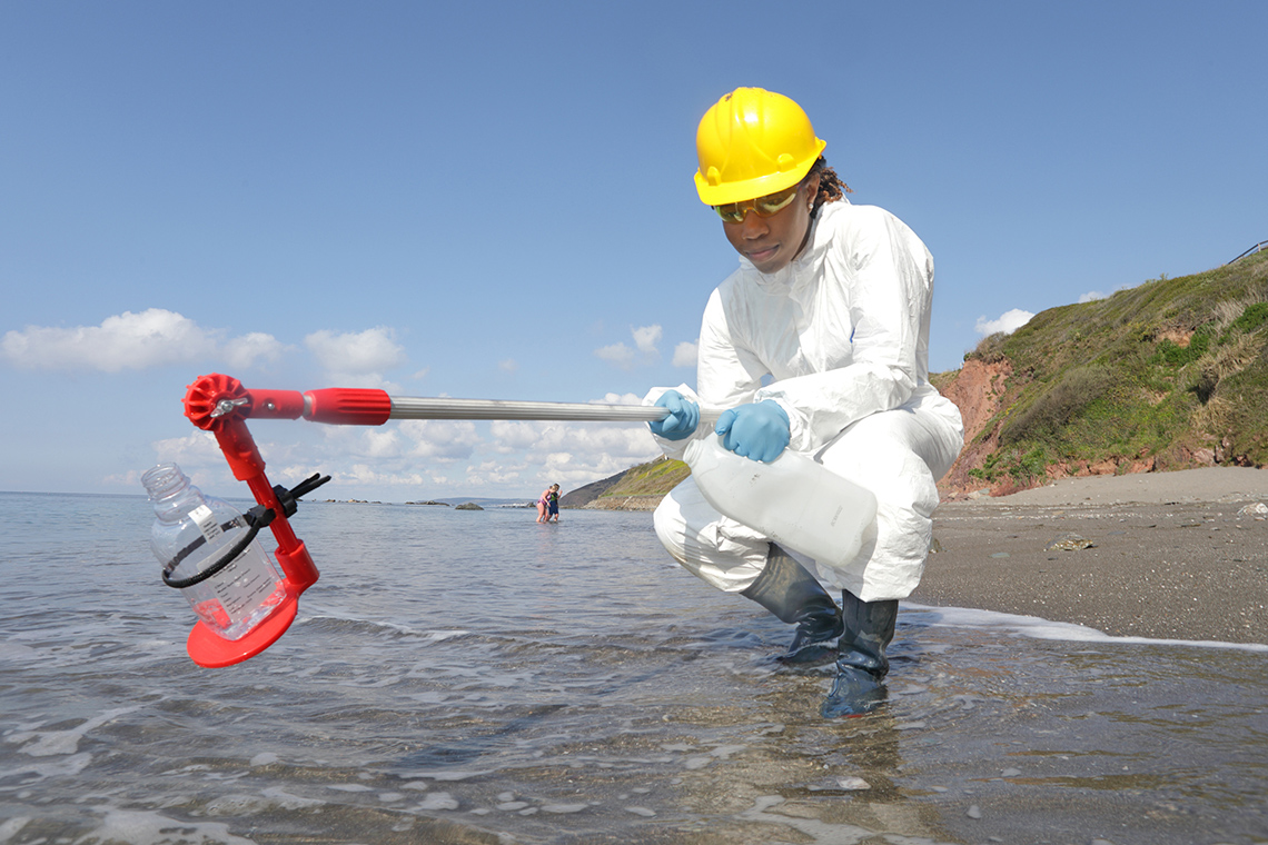 Person wearing protective equipment collecting water samples close to the shore.