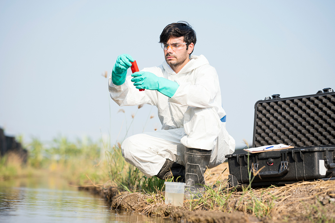 Person wearing protective equipment collecting water samples at the edge of a stream.