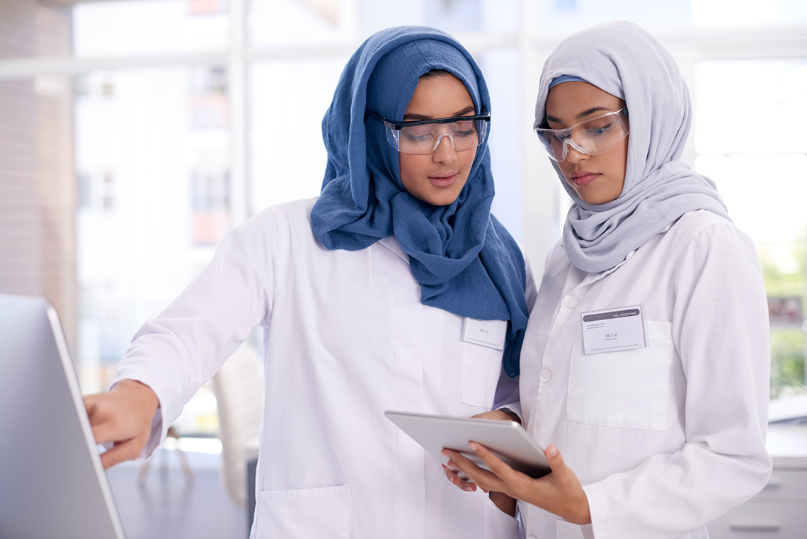 Two people wearing lab coats and safety glasses entering data into a computer.