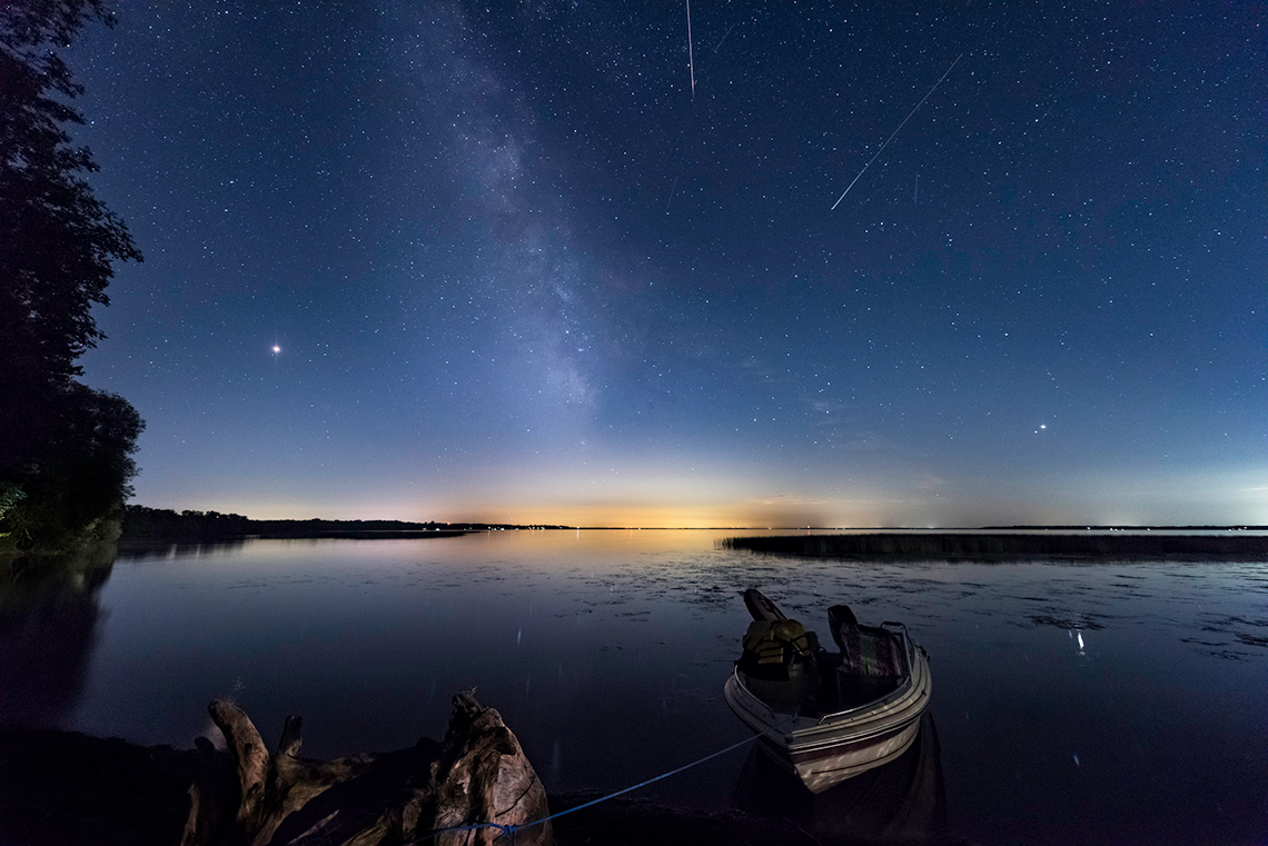 Milky Way, two meteors, and the moon in the sky above a lake at Sunset.