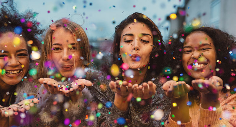 Young women blowing confetti from hands. Friends celebrating outdoors in evening at a terrace.