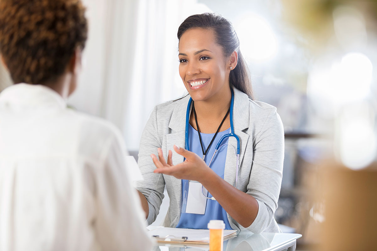 Confident mid adult African American doctor gestures while talking with an unrecognizable female patient. The doctor is reviewing the patient’s prescription medication.